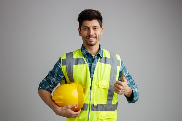 白い背景に分離された親指を上に表示しているカメラを見て安全ヘルメットを保持している制服を着て笑顔の若い男性エンジニア