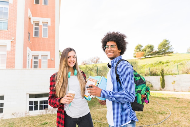 持ち帰り用のコーヒーカップと本を手で保持している若い男性と女性の学生をキャンパスで笑顔