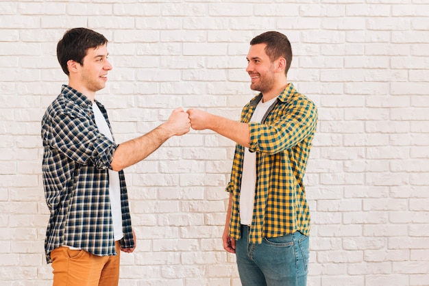 Smiling young male friends standing against white wall bumping their fist