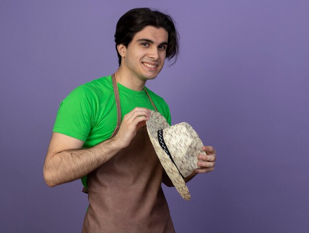 Smiling young male gardener in uniform holding gardening hat isolated on purple with copy space