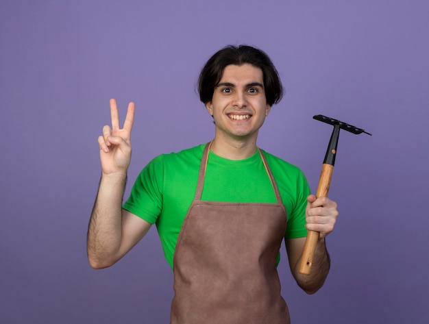 Smiling young male gardener in uniform holding hoe rake showing peace gesture