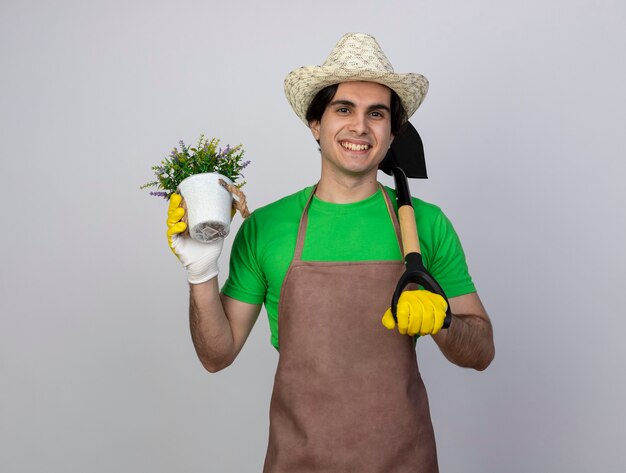 Smiling young male gardener in uniform wearing gardening hat and gloves holding flower in flowerpot and putting spade on shoulder