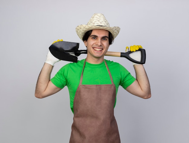 Sorridente giovane giardiniere maschio in uniforme indossando guanti e cappello da giardinaggio tenendo la vanga dietro al collo