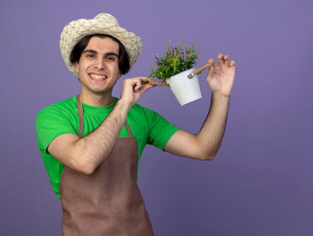 Smiling young male gardener in uniform wearing gardening hat holding flower in flowerpot isolated on purple