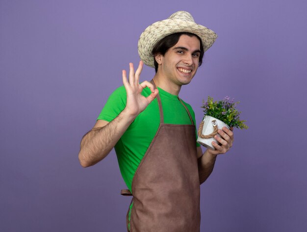 Улыбающийся молодой мужчина-садовник в униформе в садовой шляпе держит цветок в цветочном горшке, показывая хороший жест