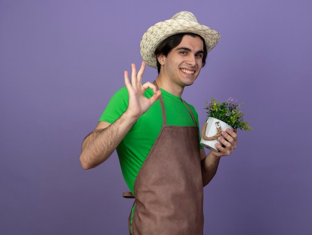 Smiling young male gardener in uniform wearing gardening hat holding flower in flowerpot showing okay gesture