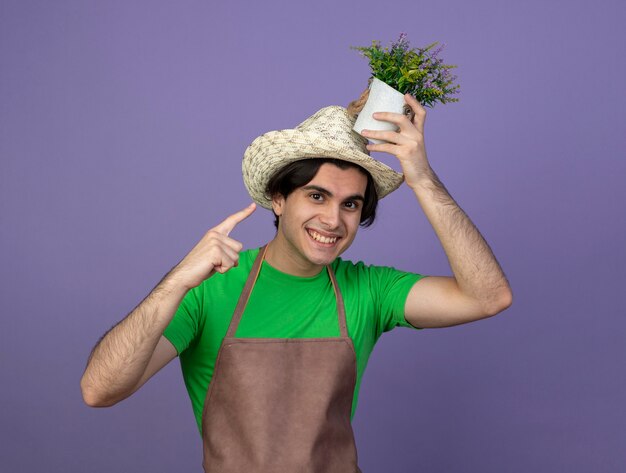 Smiling young male gardener in uniform wearing gardening hat holding and points at flower in flowerpot on head