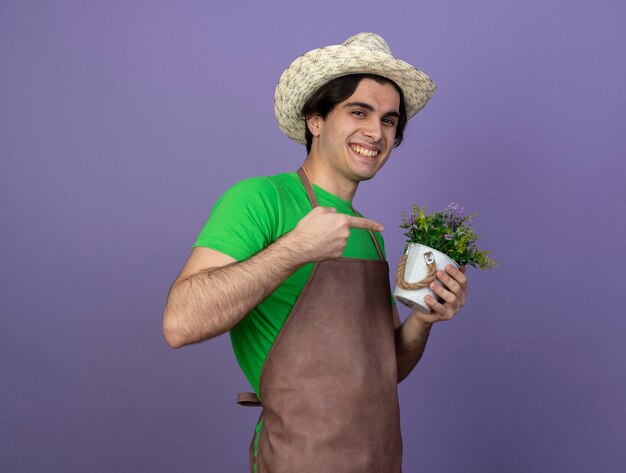 Free photo smiling young male gardener in uniform wearing gardening hat holding and points at flower in flowerpot