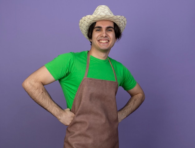 Foto gratuita sorridente giovane giardiniere maschio in uniforme che indossa il cappello da giardinaggio mettendo le mani sui fianchi isolati su viola