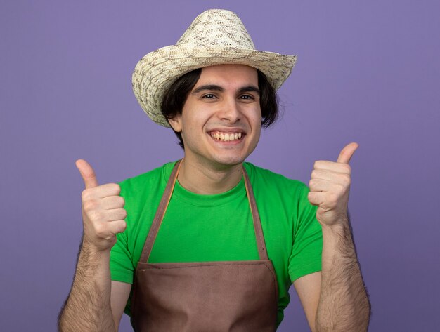 Free photo smiling young male gardener in uniform wearing gardening hat showing thumbs up isolated on purple