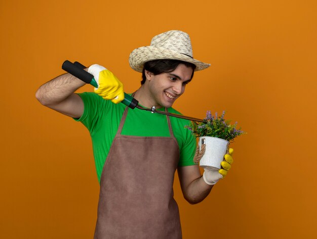 Smiling young male gardener in uniform wearing gardening hat with gloves holding clippers and cut flower in flowerpot isolated on orange