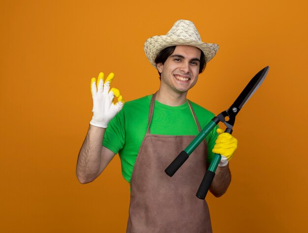 オレンジ色に分離された大丈夫なジェスチャーを示すクリッパーズを保持している手袋と園芸帽子をかぶって制服を着た若い男性の庭師の笑顔
