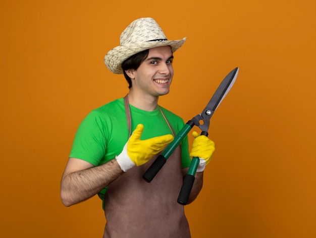 Улыбающийся молодой мужчина-садовник в униформе в садовой шляпе с перчатками держит и указывает рукой на машинки для стрижки