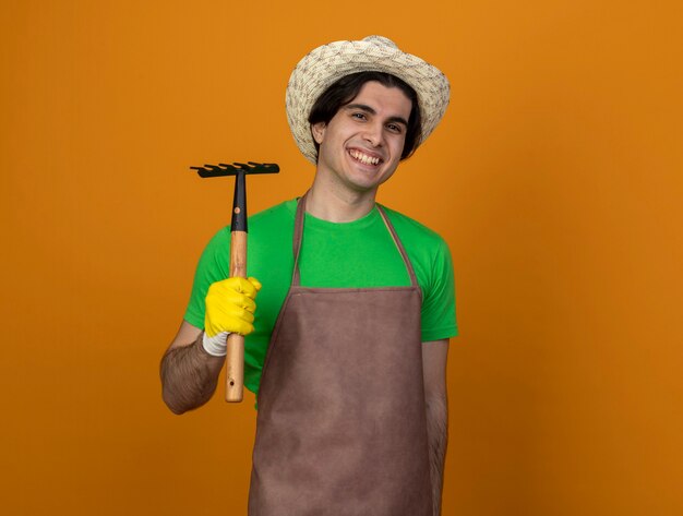 Smiling young male gardener in uniform wearing gardening hat with gloves holding rake isolated on orange with copy space