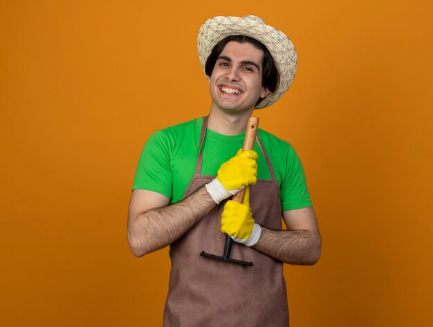 Улыбающийся молодой мужчина-садовник в униформе в садовой шляпе с перчатками держит грабли, изолированные на оранжевом
