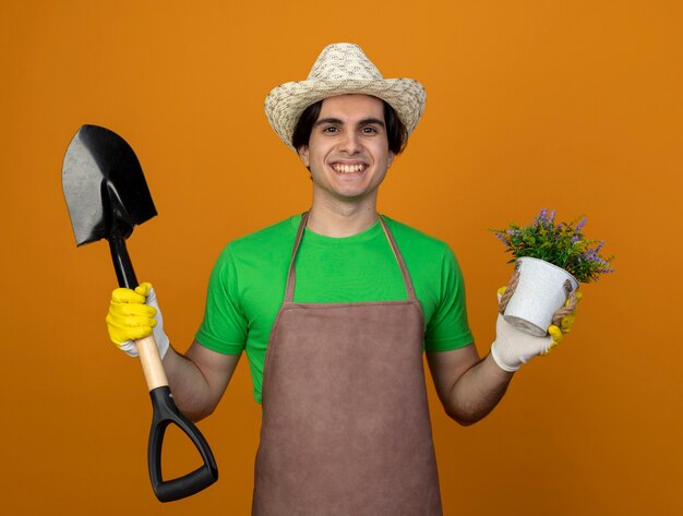 Smiling young male gardener in uniform wearing gardening hat with gloves holding spade with flower in flowerpot isolated on orange