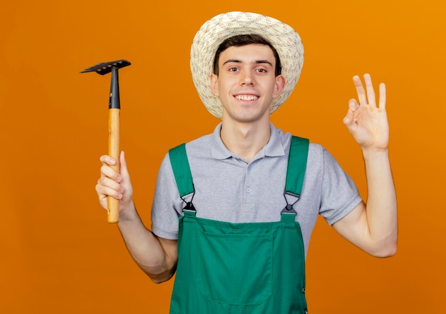 Улыбающийся молодой мужчина-садовник в садовой шляпе жестикулирует рукой и держит грабли