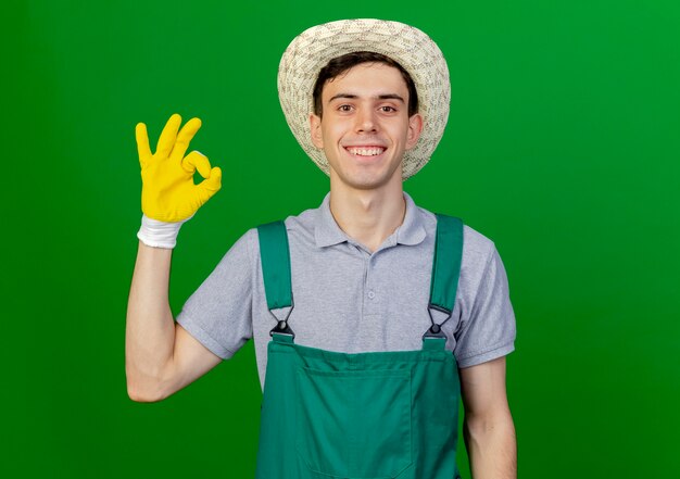 Smiling young male gardener wearing gardening hat and gloves gestures ok hand sign isolated on green background with copy space