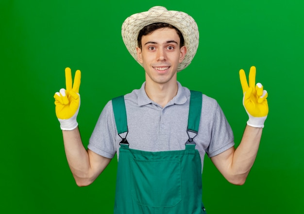 Sorridente giovane giardiniere maschio indossando guanti e cappello da giardinaggio gesti il segno della mano di vittoria con due mani isolate su priorità bassa verde con lo spazio della copia