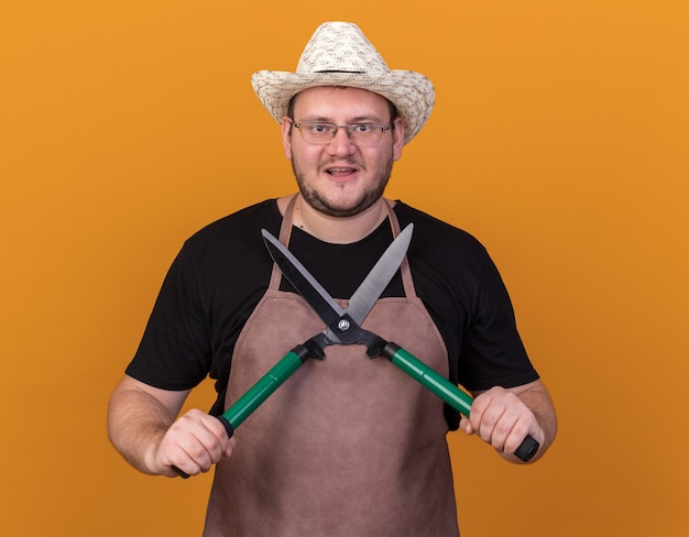 Smiling young male gardener wearing gardening hat and gloves holding clippers isolated on orange wall