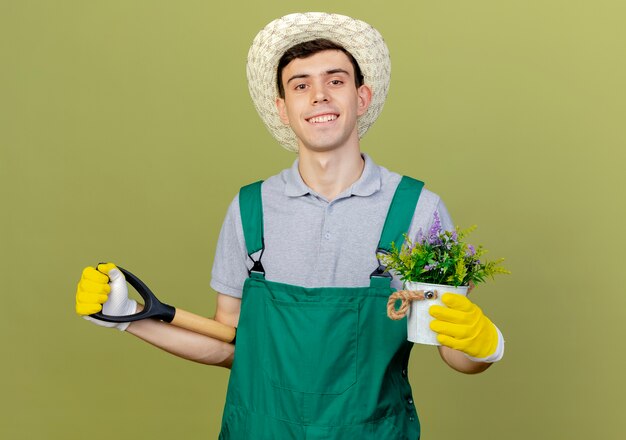 Улыбающийся молодой мужчина-садовник в садовой шляпе и перчатках держит цветы в цветочном горшке и лопату позади