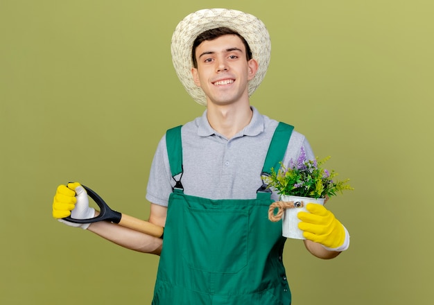 Smiling young male gardener wearing gardening hat and gloves holds flowers in flowerpot and spade behind 