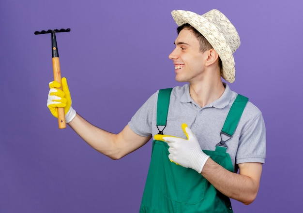 Smiling young male gardener wearing gardening hat and gloves holds and points at rake