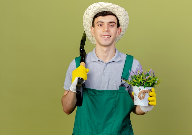 Улыбающийся молодой мужчина-садовник в садовой шляпе и перчатках держит лопату и цветы в цветочном горшке