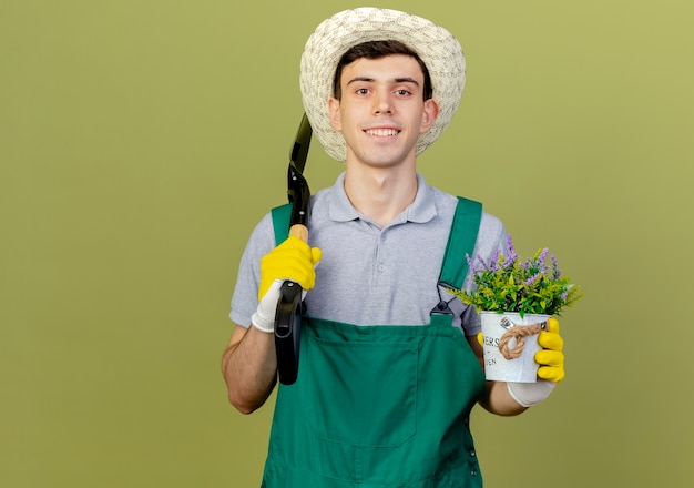 Smiling young male gardener wearing gardening hat and gloves holds spade and flowers in flowerpot 