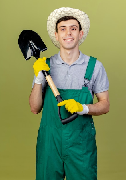 ガーデニング帽子と手袋を身に着けている笑顔の若い男性の庭師は、コピースペースでオリーブグリーンの背景に分離されたカメラを見てスペードを保持します。