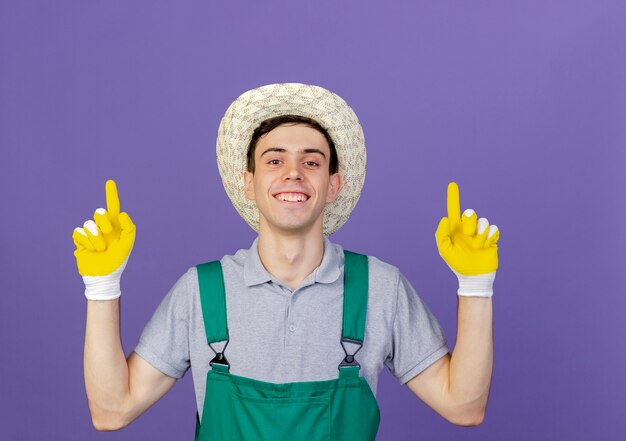 Smiling young male gardener wearing gardening hat and gloves points up