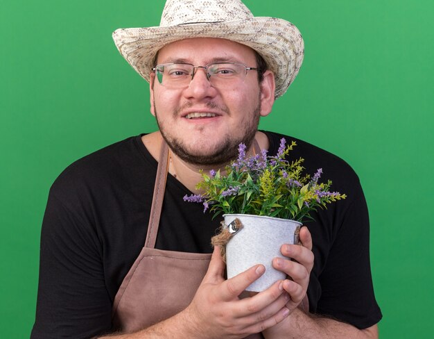 緑の壁に分離された植木鉢の花を保持しているガーデニング帽子を身に着けている若い男性の庭師の笑顔