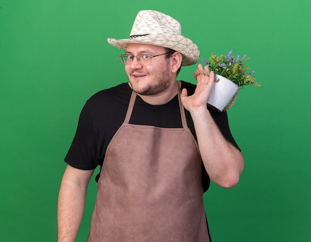 녹색 벽에 고립 된 어깨에 화분에 꽃을 들고 원예 모자를 쓰고 웃는 젊은 남성 정원사