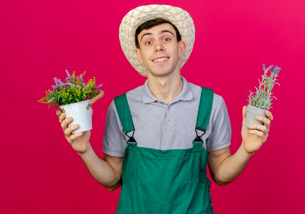 Smiling young male gardener wearing gardening hat holding flowerpots isolated on pink background with copy space