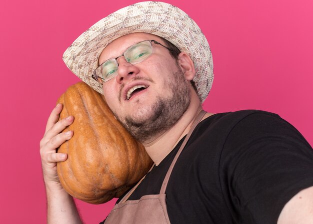 Free photo smiling young male gardener wearing gardening hat holding pumpkin and front isolated on pink wall