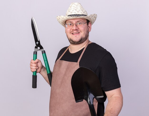 Smiling young male gardener wearing gardening hat holding spade with clippers isolated on white wall