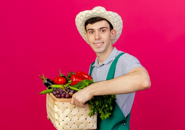 Sorridente giovane giardiniere maschio che indossa cappello da giardinaggio che tiene cesto di verdure