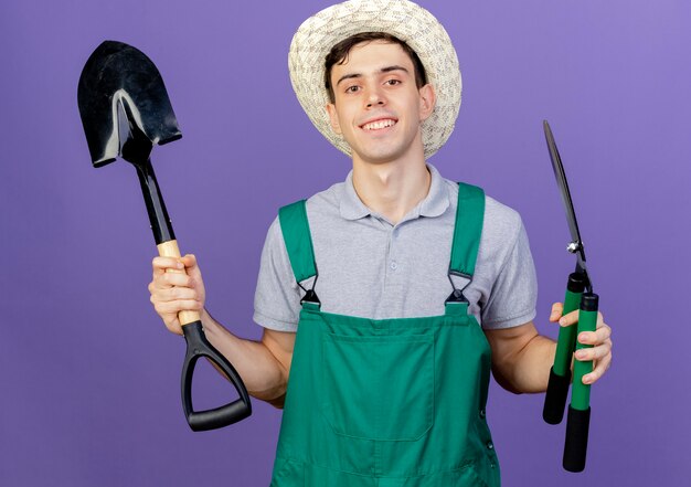 Улыбающийся молодой мужчина-садовник в садовой шляпе держит ножницы и лопату
