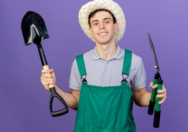 Улыбающийся молодой мужчина-садовник в садовой шляпе держит ножницы и лопату