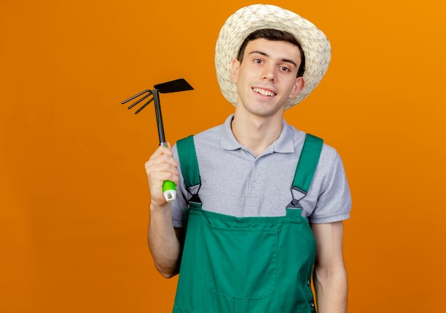 Smiling young male gardener wearing gardening hat holds hoe rake 