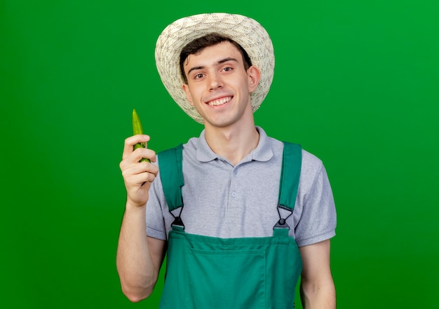 Smiling young male gardener wearing gardening hat holds hot pepper isolated on green background with copy space