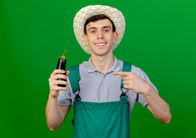 Smiling young male gardener wearing gardening hat holds and points at eggplant isolated on green background with copy space