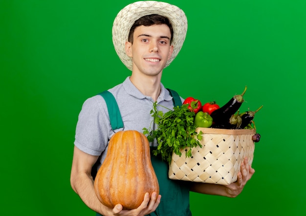 Бесплатное фото Улыбающийся молодой мужчина-садовник в садовой шляпе держит корзину с тыквами и овощами