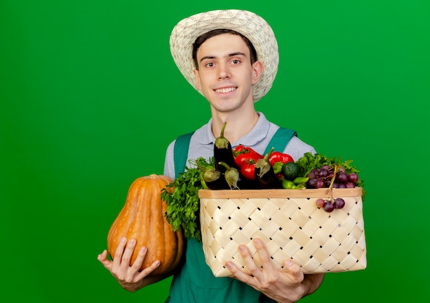 Улыбающийся молодой мужчина-садовник в садовой шляпе держит корзину с тыквами и овощами