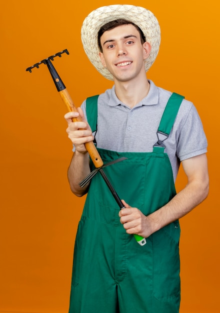 Free photo smiling young male gardener wearing gardening hat holds rake over hoe rake isolated on orange background with copy space