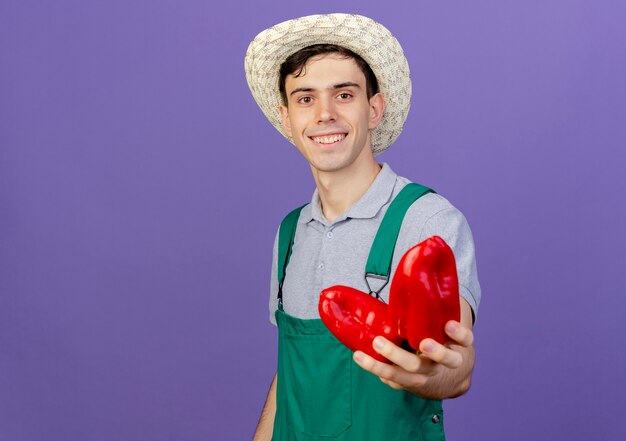 Smiling young male gardener wearing gardening hat holds red peppers