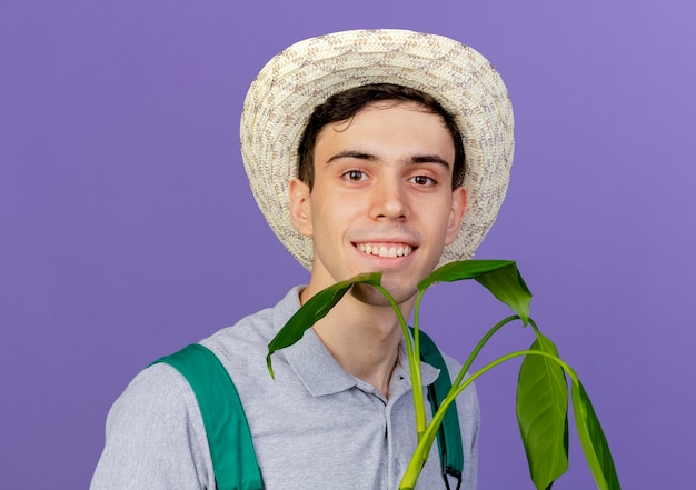 Smiling young male gardener wearing gardening hat stands with plant 