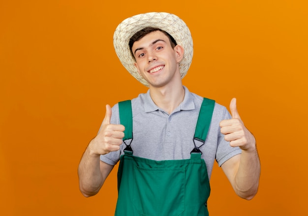 Foto gratuita sorridente giovane giardiniere maschio che indossa il cappello da giardinaggio pollice in alto con due mani