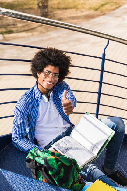 Бесплатное фото Улыбающийся молодой ученик, сидя на лестнице, показывая большой палец вверх знак