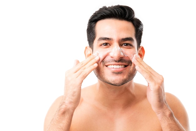 Smiling young man applying anti-aging cream on cheeks while making eye contact against white background